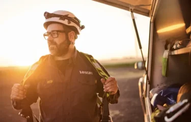 A Vestas technician wearing safety gear and a helmet stands beside a utility vehicle at sunrise, adjusting his harness while looking into the distance. The open vehicle door reveals work equipment inside.